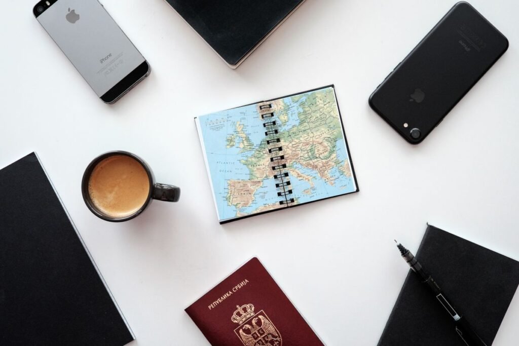 Flat lay of two smartphones, a European map notebook, a passport, and a coffee cup on a white desk, representing the groundwork of organising local business information